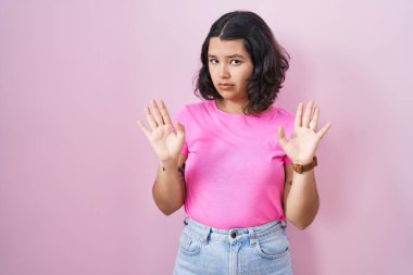 Young hispanic woman standing over pink background moving away hands palms showing refusal and denial with afraid and disgusting expression. stop and forbidden. 