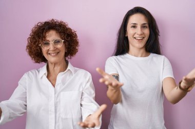 İspanyol anne ve kızı pembe arka plan üzerine günlük beyaz tişört giyip neşeli gülümseyerek yardım eli uzatıyor ve kabul görüyor.. 