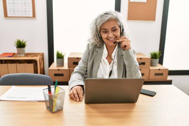 Orta yaşlı, gri saçlı çağrı merkezi ajanı kadın ofiste çalışırken mutlu bir şekilde gülümsüyor..