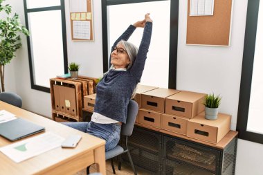 Orta yaşlı, kır saçlı, iş adamı bir kadın ofiste kollarını açmış dinleniyor.