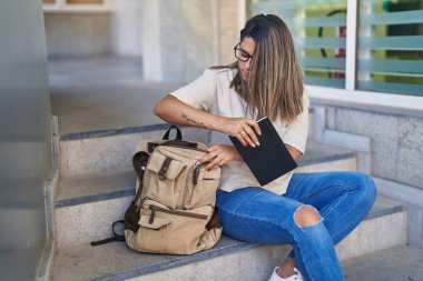 Young hispanic woman student putting notebook and book on backpack at university