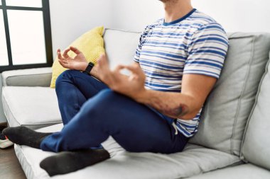 Genç İspanyol, yoga egzersizi yapıyor. Evdeki kanepede oturuyor.