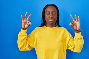 Beautiful black woman standing over blue background relaxed and smiling with eyes closed doing meditation gesture with fingers. yoga concept. 