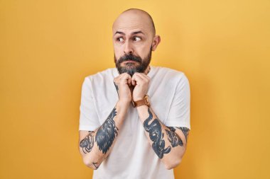 Sarı arka planda dikilen dövmeleri olan genç İspanyol adam elleri çenesinde gergin ve heyecanlı bir şekilde yan tarafa bakıyor. 