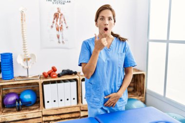 Middle age hispanic physiotherapist woman working at pain recovery clinic surprised pointing with finger to the side, open mouth amazed expression. 