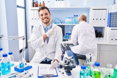 Hispanic man and woman working at scientist laboratory smiling happy pointing with hand and finger to the side 