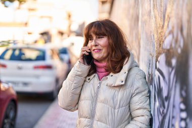Orta yaşlı bir kadın gülümsüyor. Sokakta akıllı telefondan konuşuyor.