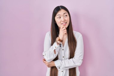 Çinli genç bir kadın pembe arka planda, elleri çenesinde soru sorarken, düşünceli bir ifade takınıyor. Gülümseyen ve düşünceli bir yüz. şüphe kavramı. 