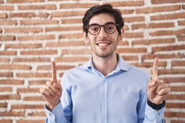 Young hispanic man standing over brick wall background smiling amazed and surprised and pointing up with fingers and raised arms. 