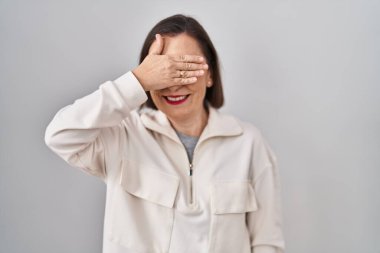 Orta yaşlı İspanyol bir kadın izole edilmiş arka planda duruyor. Gülümsüyor ve gülümsüyor. Gözlerini şaşkınlıkla kapatıyor. kör kavram. 