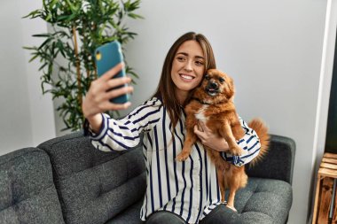 Genç İspanyol kadın akıllı telefonun başında selfie çekiyor. Kanepede oturup evdeki köpeğe sarılıyor.