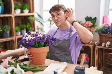 Caucasian blond man working at florist shop smiling with hand over ear listening an hearing to rumor or gossip. deafness concept. 
