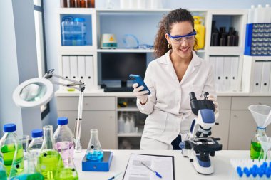 Young beautiful hispanic woman scientist smiling confident using smartphone at laboratory