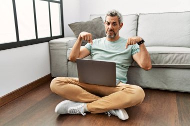 Orta yaşlı İspanyol adam oturma odasında dizüstü bilgisayar kullanıyor üzgün ve üzgün görünüyor, parmaklarıyla yönü gösteriyor, mutsuz ve depresif.. 