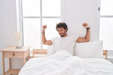 Young hispanic man waking up stretching arms at bedroom