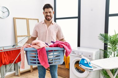 Young man with beard holding laundry basket sticking tongue out happy with funny expression. 