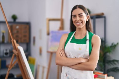 Genç İspanyol kadın sanatçı gülümsüyor. Kollarını kavuşturup sanat stüdyosunda duruyor.
