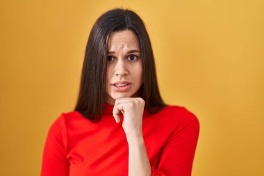 Sarı arka planda dikilen genç İspanyol kadın gergin ve gergin görünüyor. Elleri ağızda, tırnaklarını yiyormuş. anksiyete. 