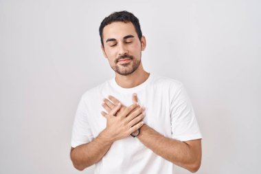 Handsome hispanic man standing over white background smiling with hands on chest with closed eyes and grateful gesture on face. health concept. 