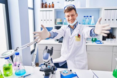 Caucasian man working at scientist laboratory looking at the camera smiling with open arms for hug. cheerful expression embracing happiness. 