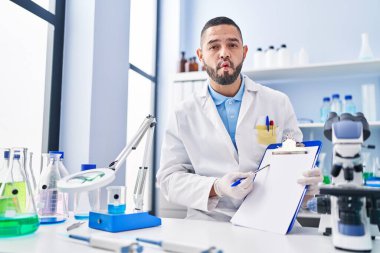 Hispanic man working at scientist laboratory holding blank clipboard making fish face with mouth and squinting eyes, crazy and comical. 