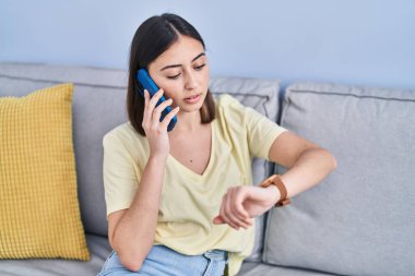Young hispanic woman talking on the smartphone looking watch at home