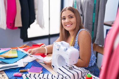 Young beautiful hispanic woman tailor smiling confident holding clothing design at tailor shop