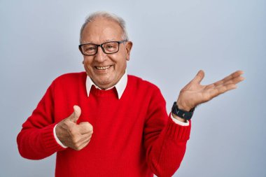Gri saçlı yaşlı adam arka planda duruyor. Avuç içlerini gösteriyor ve baş parmaklarıyla iyi bir hareket yapıyor. Mutlu ve neşeli gülümsüyor. 
