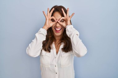 Hispanic woman standing over isolated background doing ok gesture like binoculars sticking tongue out, eyes looking through fingers. crazy expression. 