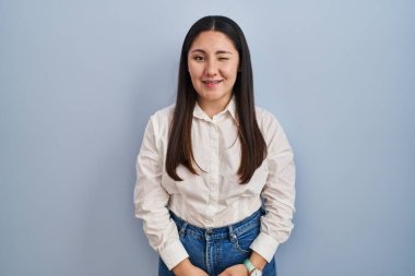 Young latin woman standing over blue background winking looking at the camera with sexy expression, cheerful and happy face. 