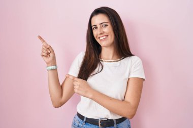 Young brunette woman standing over pink background smiling and looking at the camera pointing with two hands and fingers to the side. 