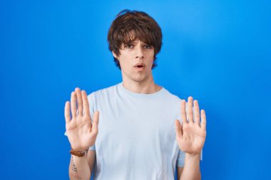 İspanyol genç adam mavi arka planda duruyor. Avuçlarını hareket ettiriyor. Korkmuş ve iğrenç bir ifadeyle reddediyor. Dur ve yasakla. 