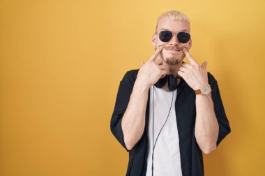 Sarı arka planda duran güneş gözlüklü genç beyaz adam ağzı açık gülümsüyor, parmakları işaret ediyor ve neşeli bir gülümseme takınıyor. 