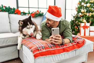 Evdeki Noel ağacında akıllı telefon kullanan genç bir İspanyol adam köpekle kanepede yatıyor.