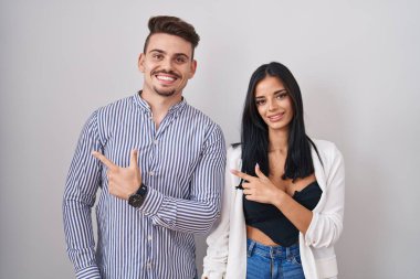 Young hispanic couple standing over white background cheerful with a smile on face pointing with hand and finger up to the side with happy and natural expression 