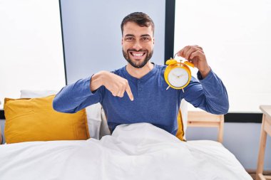 Handsome hispanic man in the bed holding alarm clock pointing finger to one self smiling happy and proud 