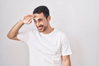 Handsome hispanic man standing over white background very happy and smiling looking far away with hand over head. searching concept. 