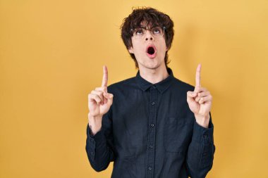 Sarı arka planda gözlüklü genç adam hayret ve şaşkınlık içinde yukarı baktı ve parmaklarıyla işaret ederek kollarını kaldırdı.. 