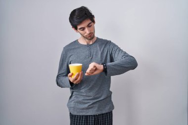 Young hispanic man wearing pajama drinking a cup of coffee checking the time on wrist watch, relaxed and confident 