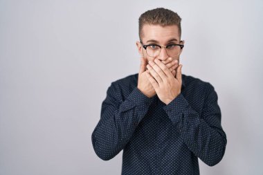 Young caucasian man standing over isolated background shocked covering mouth with hands for mistake. secret concept. 