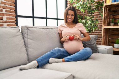 Kalbini taşıyan hamile genç Latin kadın evdeki kanepede oturuyor.