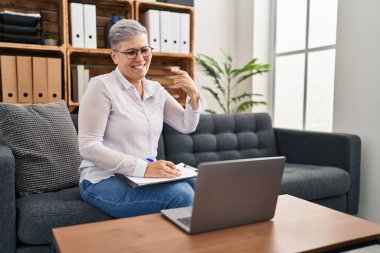 Orta yaşlı bir kadın danışma ofisinde online seans yapıyor mutlu bir şekilde el ve parmak işareti yapıyor. 