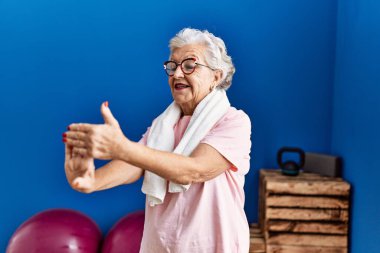 Spor kıyafetleri giyen yaşlı gri saçlı kadın spor merkezinde kendine gülerek duruyor.