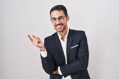 Handsome business hispanic man standing over white background with a big smile on face, pointing with hand finger to the side looking at the camera. 