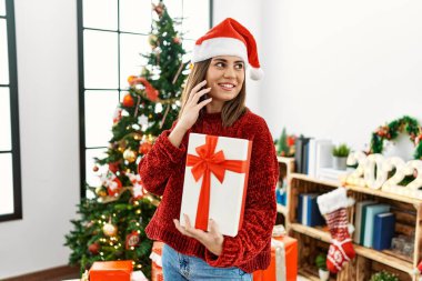 Genç İspanyol kız akıllı telefondan konuşuyor ve elinde hediyesiyle evde Noel ağacının yanında bekliyor..
