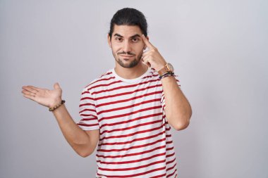 Hispanic man with long hair standing over isolated background confused and annoyed with open palm showing copy space and pointing finger to forehead. think about it. 