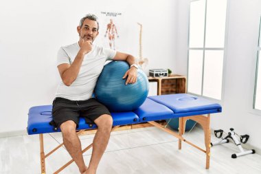Orta yaşlı İspanyol bir adam ağrı kesici kliniğinde pilates topu tutuyor kameraya kendinden emin bakıyor kollarını kavuşturmuş ve çenesini kaldırmış gülümsüyor. Olumlu düşünmek. 