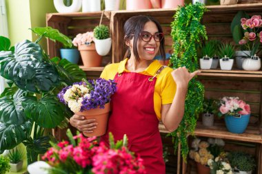 Young arab woman working at florist shop holding plant pointing thumb up to the side smiling happy with open mouth 