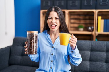 Young brunette woman holding coffee beans and cup of coffee angry and mad screaming frustrated and furious, shouting with anger. rage and aggressive concept. 