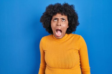 Black woman with curly hair standing over blue background angry and mad screaming frustrated and furious, shouting with anger. rage and aggressive concept. 
